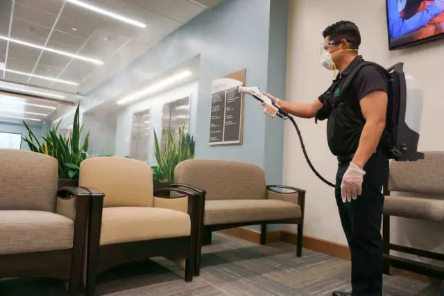 Technician in a mask disinfecting an office waiting area with an electrostatic sprayer